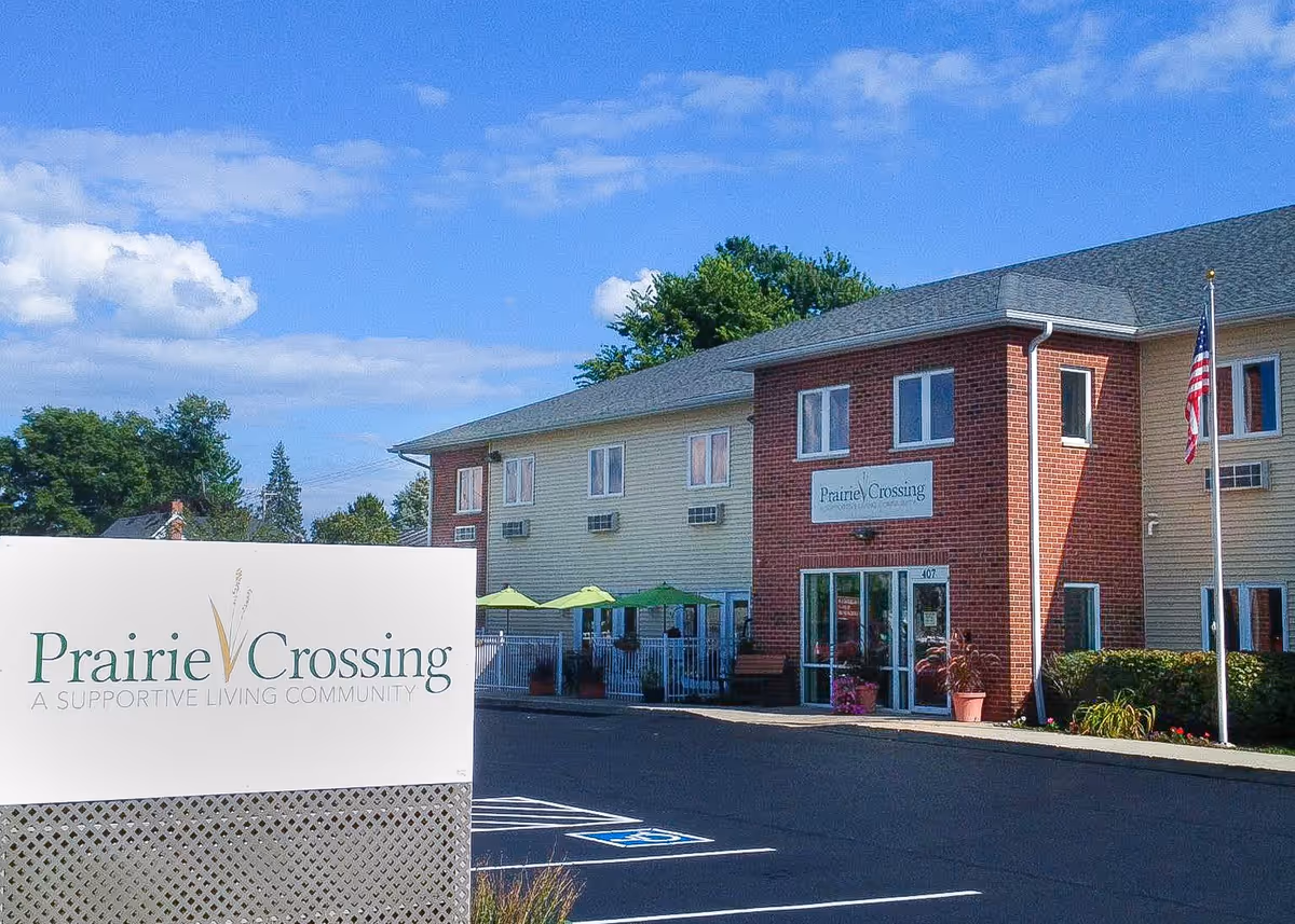 Exterior view of Prairie Crossing Assisted Living facility showing a two-story building with beige siding and red brick accents. There is a sign in the foreground with the facility name and tagline 'A Supportive Living Community'. The building has several windows, a glass entrance door, green umbrellas on a patio area, and an American flag on a flagpole. The sky is blue with some clouds.