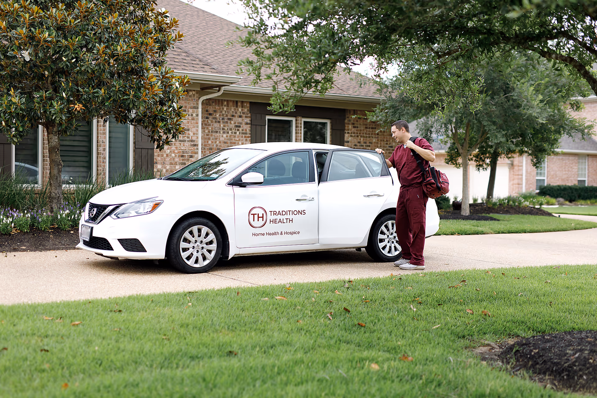 Staff member in burgundy scrubs stands beside a white hospice car in front of brick building.