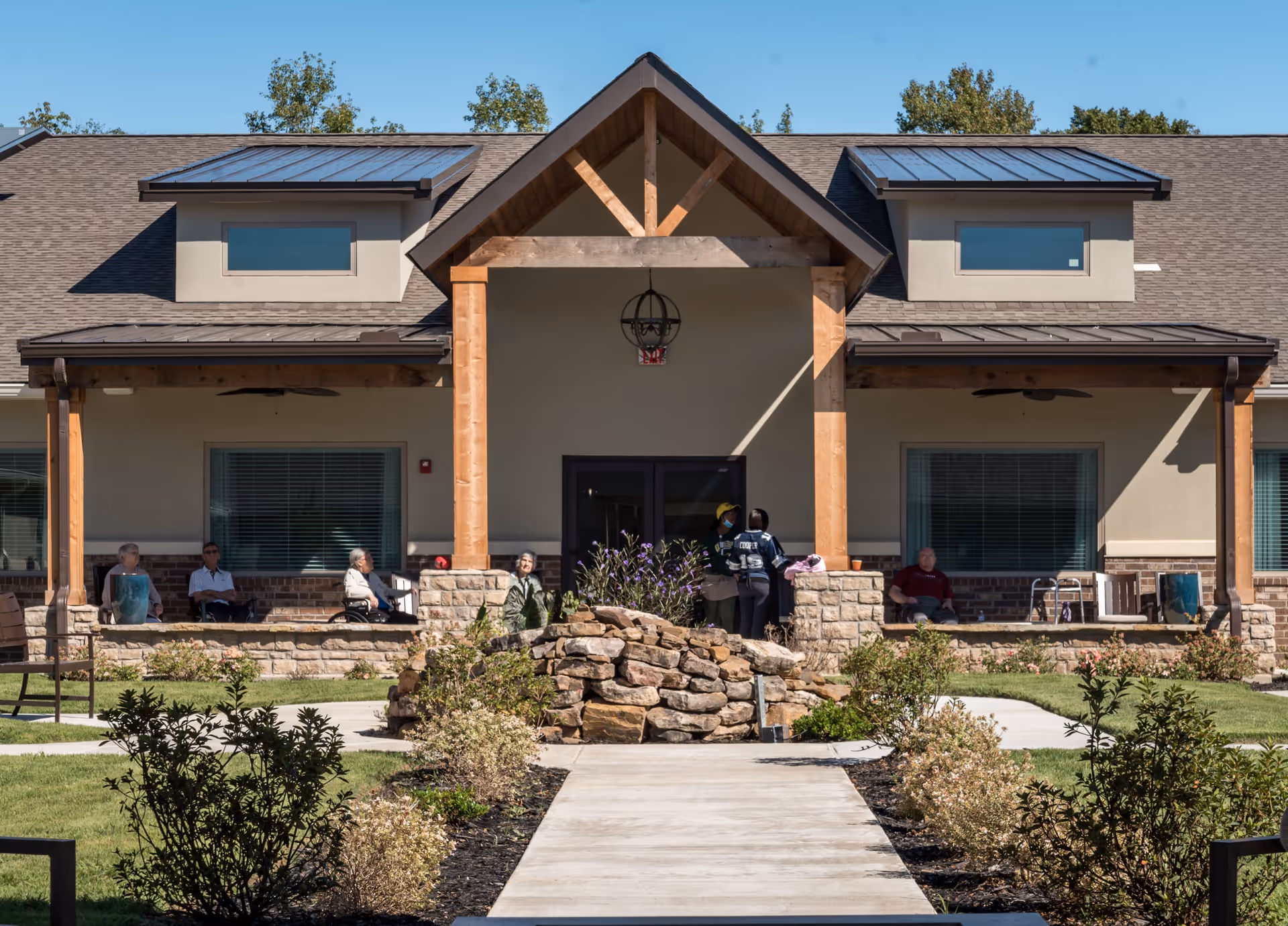 Front exterior view of Crestview Senior Living building with a covered entrance supported by wooden beams. Several elderly people are sitting on benches along the porch, and two staff members are standing near the entrance. There is a stone rock feature with plants in front of the walkway leading to the entrance. The sky is clear and blue.