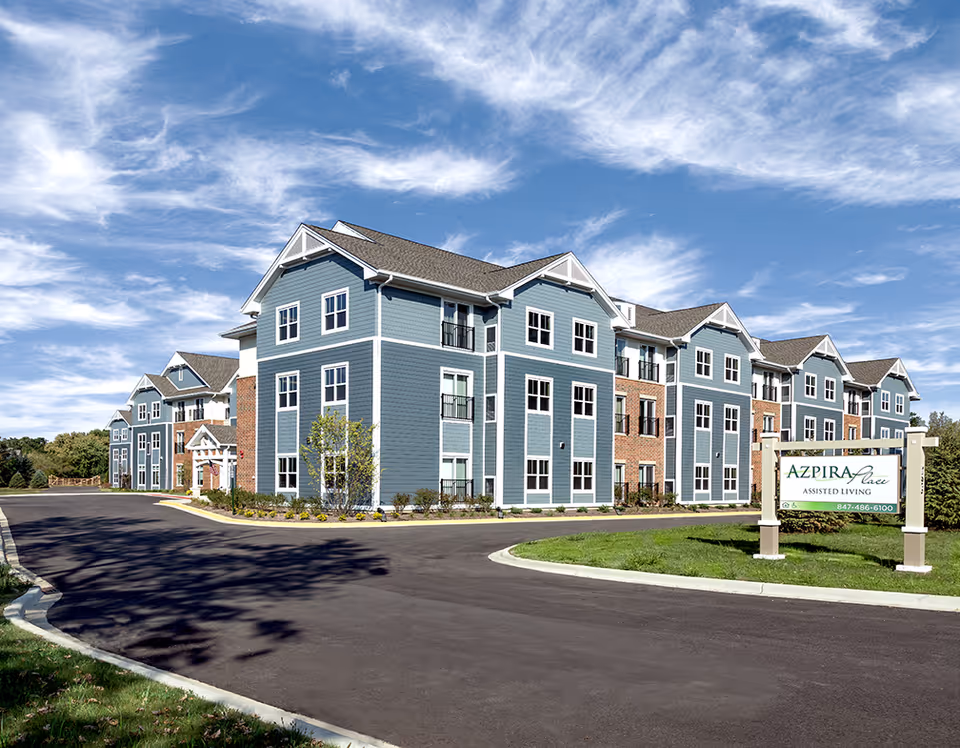 Exterior view of a large, modern assisted living building with blue siding and brick accents under a partly cloudy sky. A paved driveway curves in front of the building, and a sign in the foreground reads 'Azpira Place Assisted Living' with a phone number.