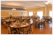Spacious dining room with multiple round wooden tables and chairs arranged neatly. Each table is set with cups, plates, and utensils. The room has large windows allowing natural light to brighten the space, with warm beige walls and ceiling lights providing additional illumination.
