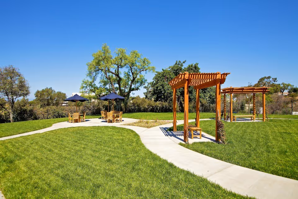 Sunlit landscaped lawn with concrete pathways, wooden pergolas and outdoor tables with umbrellas under a clear blue sky.