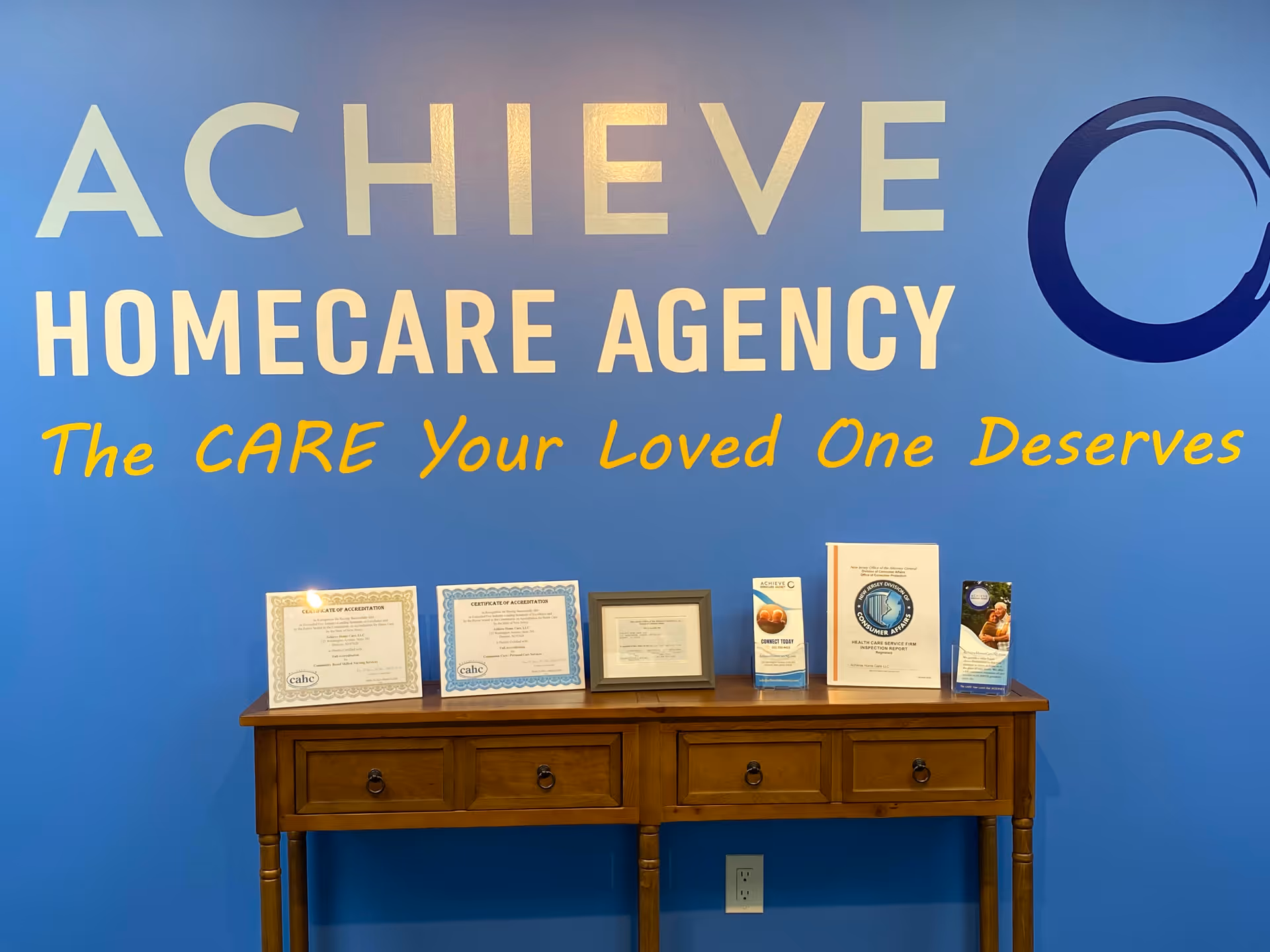 Blue wall with ACHIEVE HOMECARE AGENCY branding and a wooden console with framed certificates.