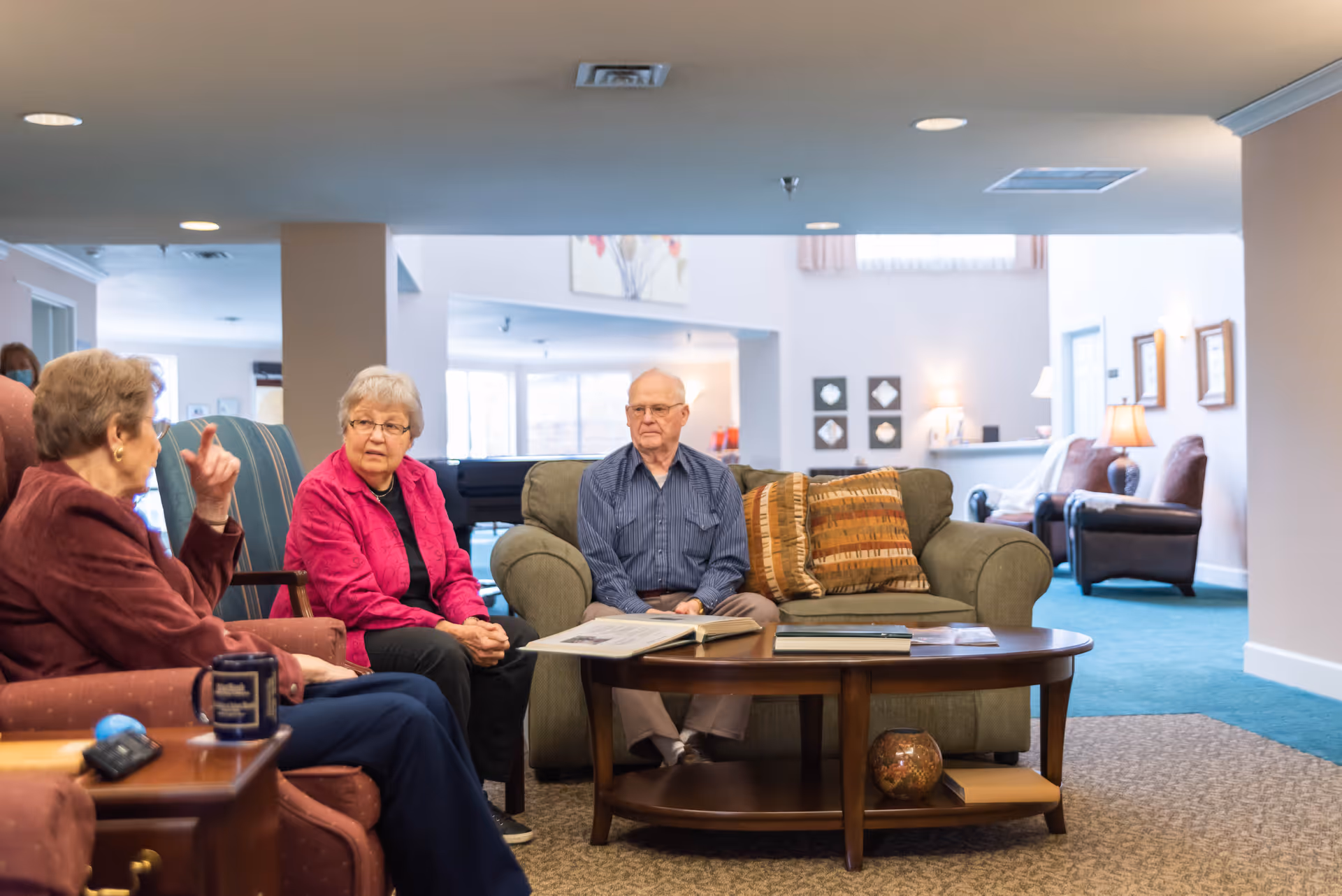 Three residents chat in a well-lit senior living facility lounge.