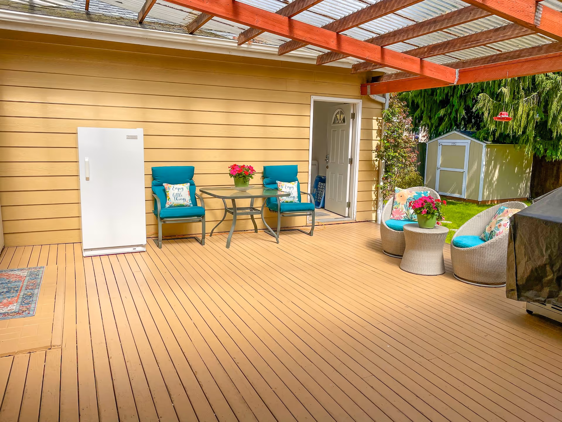 A covered outdoor patio area with wooden flooring and a transparent roof supported by wooden beams. The patio features two turquoise cushioned chairs with floral pillows and a glass-top table with a potted plant. To the right, there are two round wicker chairs with turquoise cushions and floral pillows, along with a small matching table holding another potted plant. A white door leads inside the building, and a small garden shed and greenery are visible in the background.