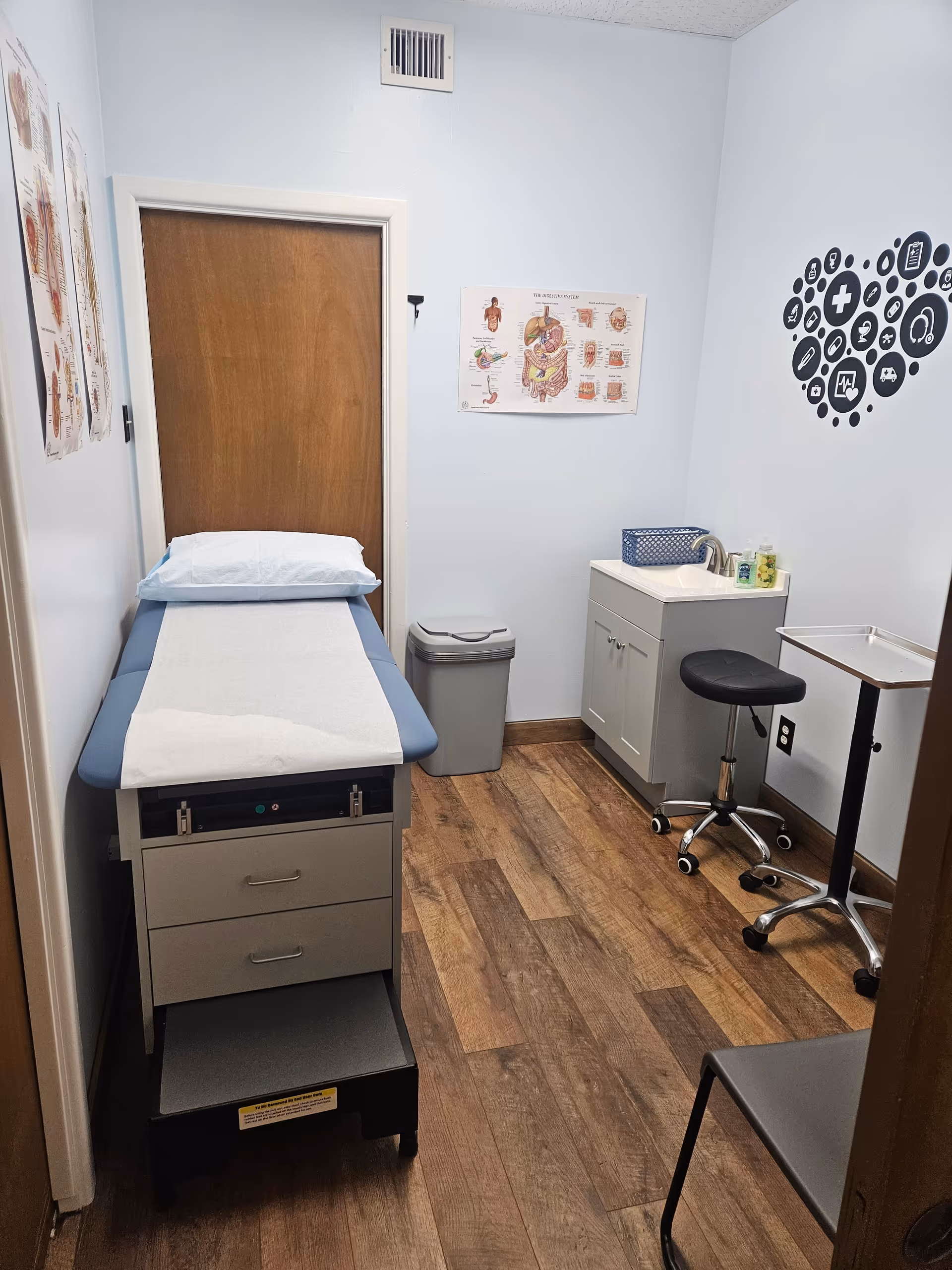 Compact medical examination room with a treatment table, sink, stool, and posters.