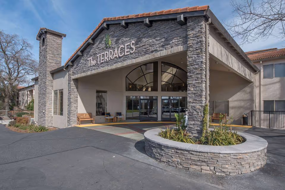 Stone-facade entrance of The Terraces senior living, with circular planter and glass doors.