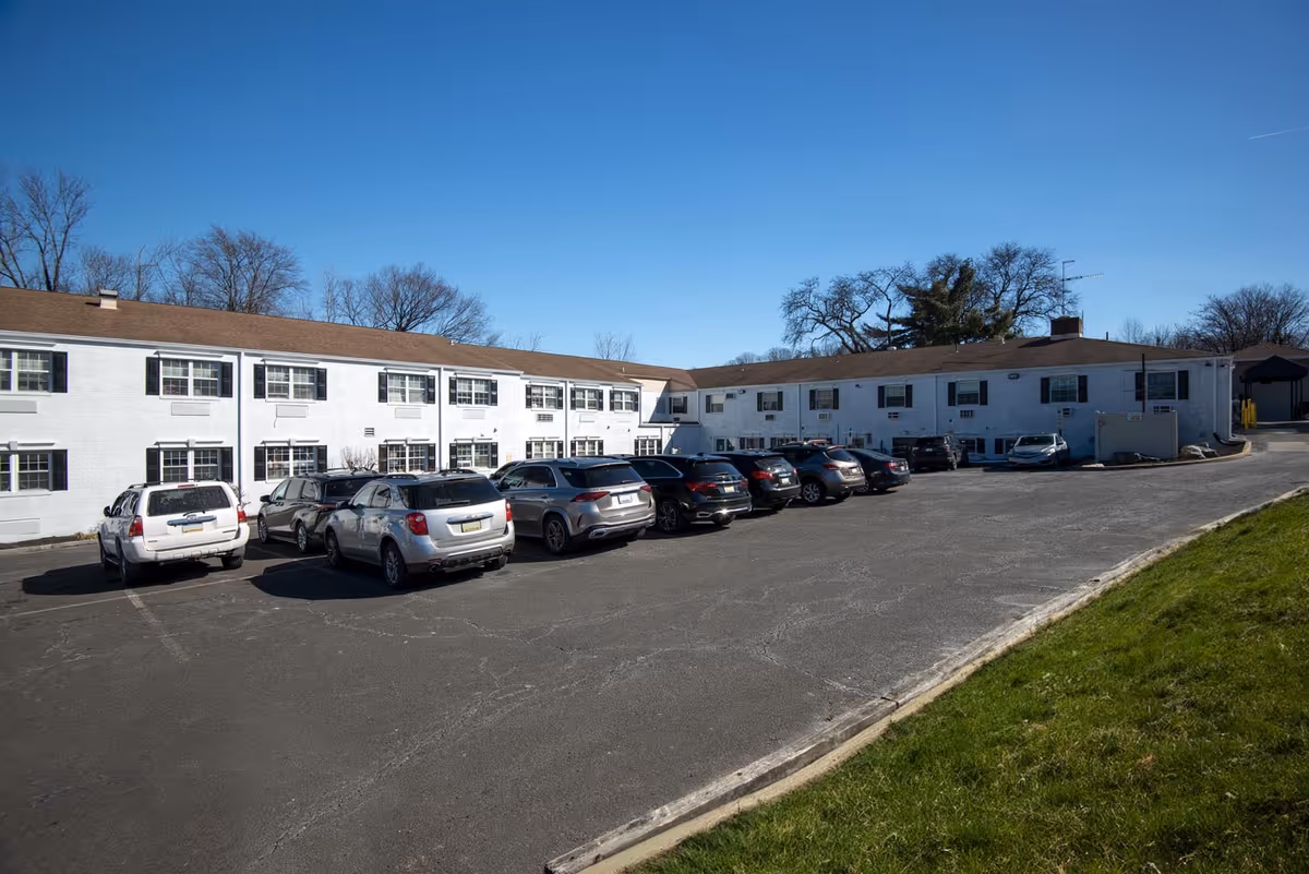 Long white two-story senior living building with a paved parking lot and blue sky.