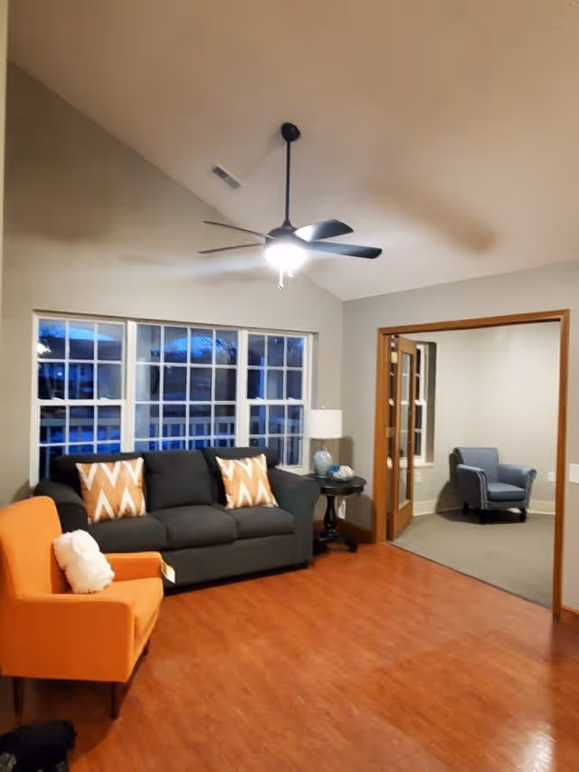 A living room with a dark gray sofa adorned with two orange and white patterned pillows, an orange armchair with a white pillow, a round side table with a blue lamp, and a ceiling fan with a light. The room has large windows and wooden flooring. An adjacent room with a blue armchair is visible through an open doorway with wooden trim.