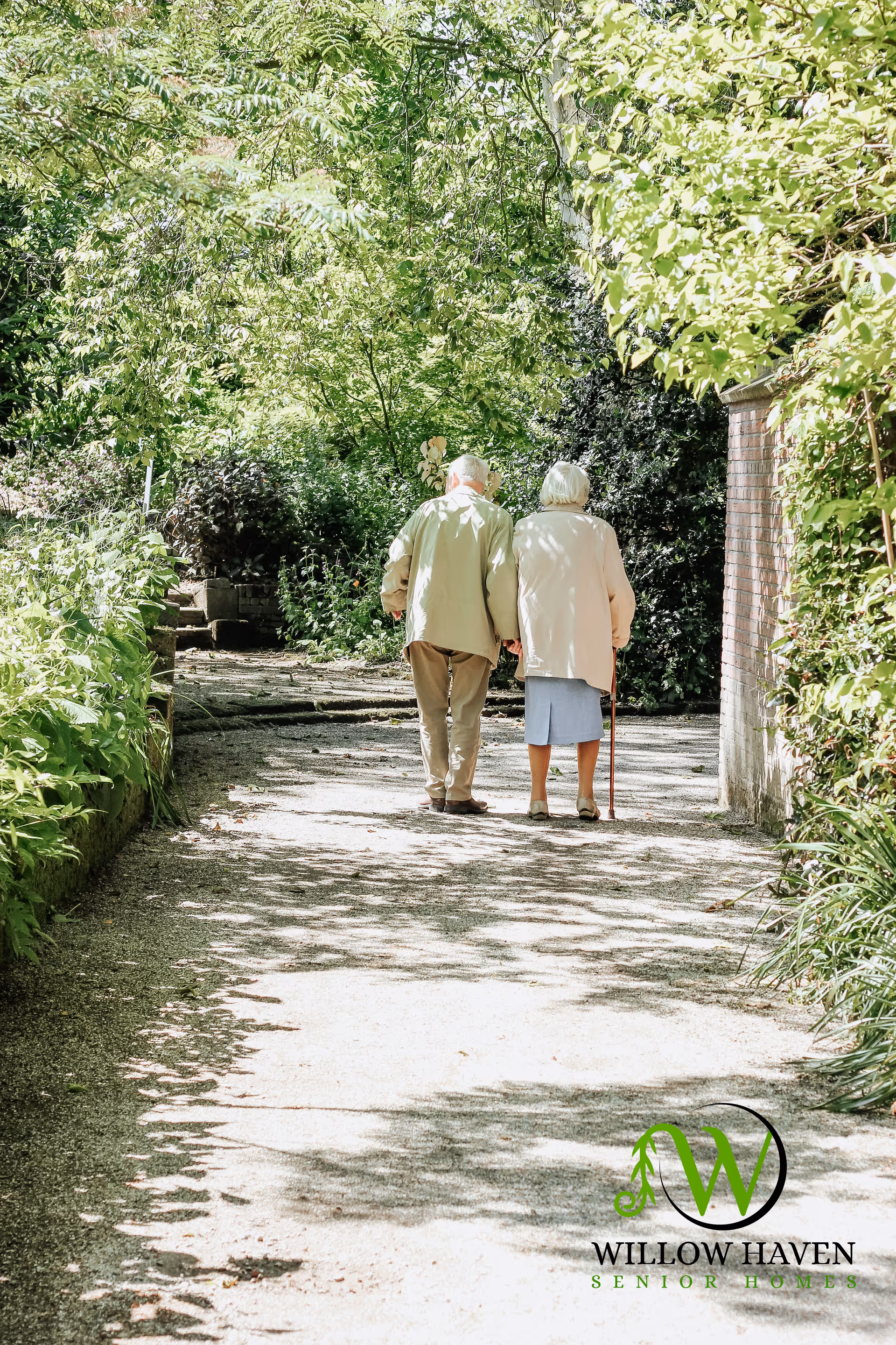 An elderly couple walking hand in hand down a shaded garden path surrounded by lush green trees and plants. The woman is using a cane and both are dressed in light-colored clothing.