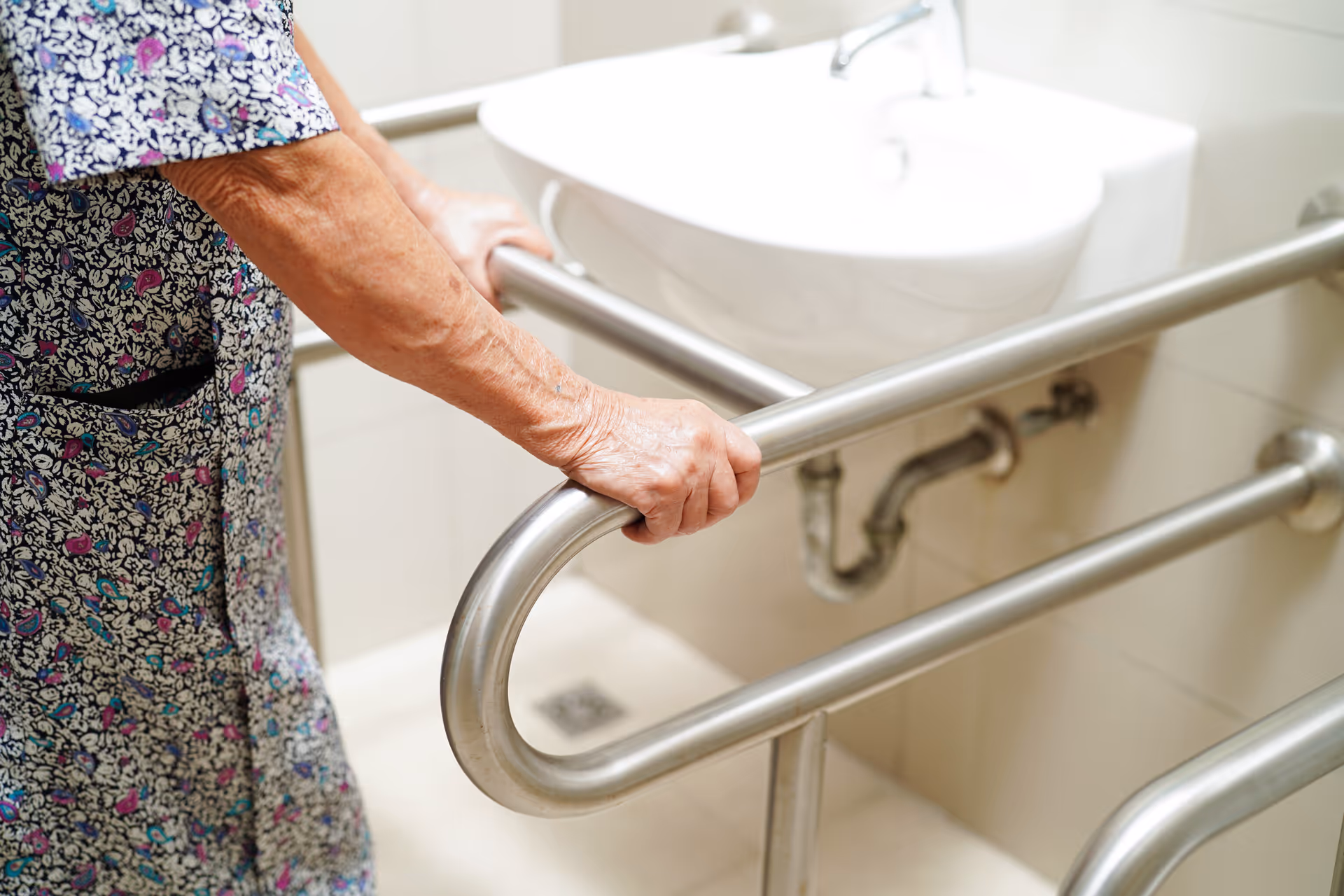 An elderly person grips a metal bathroom grab bar beside a sink.