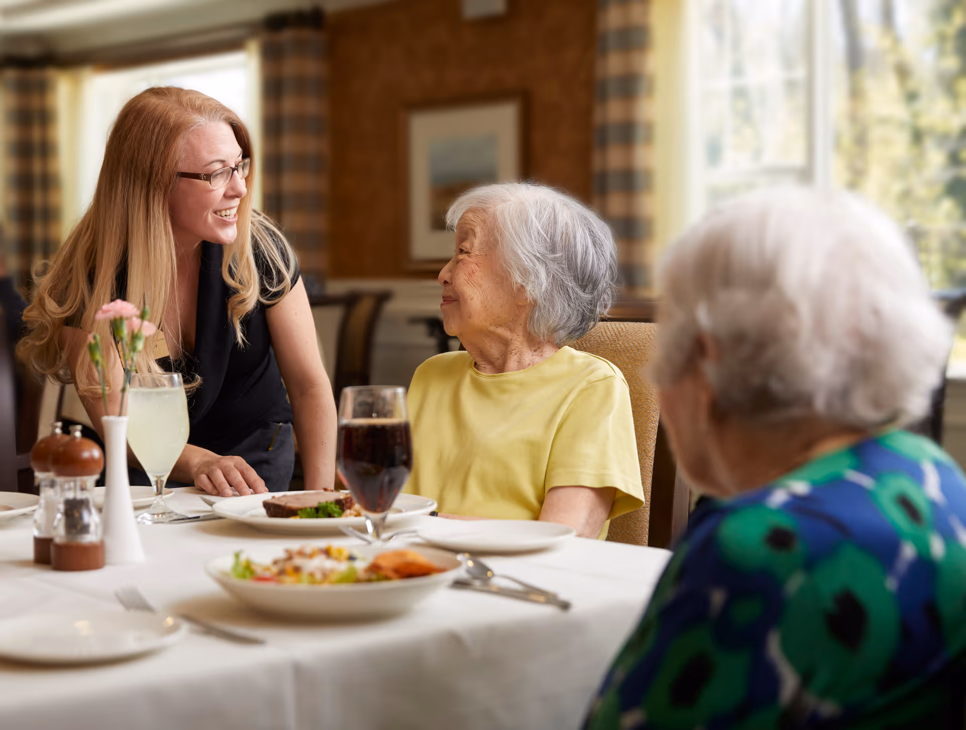 A smiling woman with long blonde hair and glasses leans toward an elderly woman with short gray hair wearing a yellow shirt, seated at a dining table with plates of food and drinks. Another elderly woman with white hair and a blue patterned top is also seated at the table. The setting appears to be a warm, well-lit dining room with large windows and curtains.