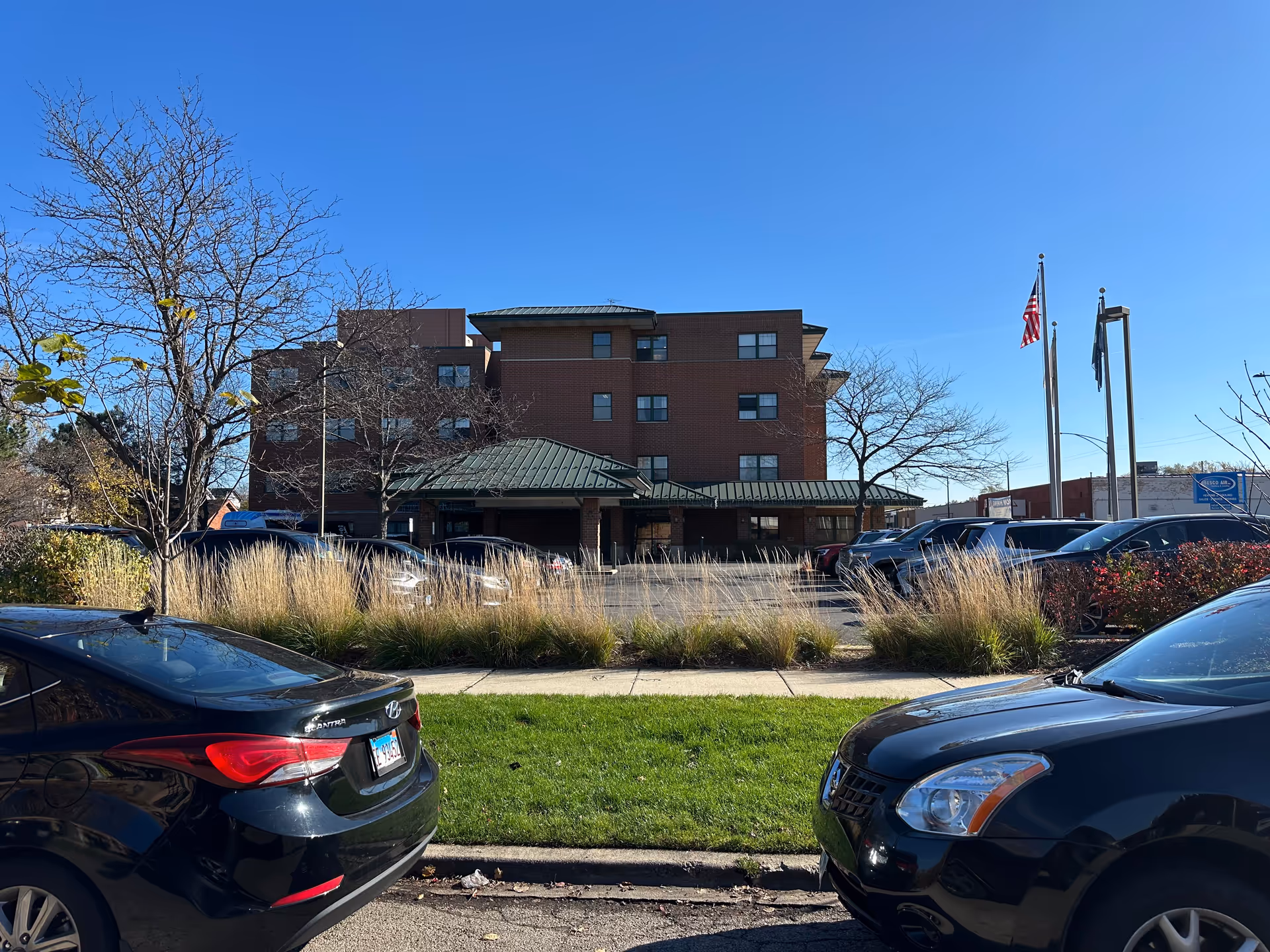 Exterior view of a multi-story brick building with a green metal roof, surrounded by parked cars and landscaping including grass, bushes, and leafless trees under a clear blue sky. Three flagpoles with flags are visible to the right of the building.