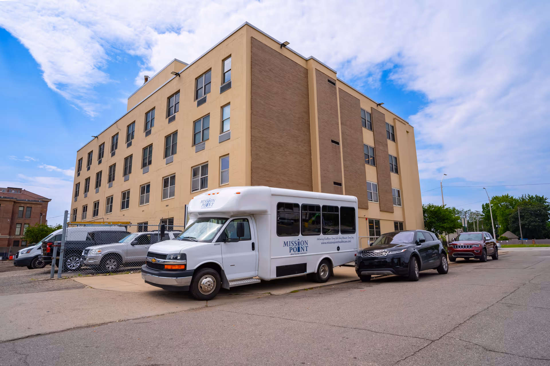 Exterior view of a multi-story beige and brown building with several windows. In front of the building, there is a white shuttle bus with the text 'MISSION POINT' parked alongside several other vehicles in a parking area. The sky is partly cloudy with blue patches.