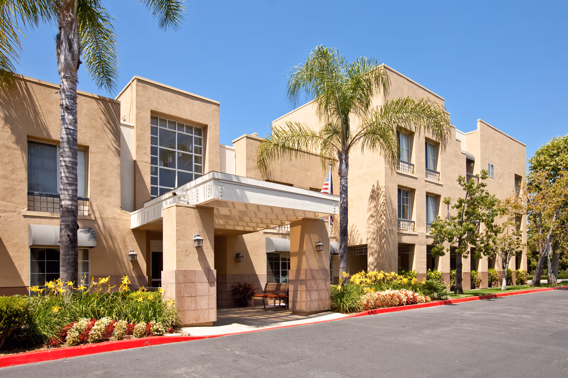 Exterior view of a beige multi-story senior living facility with palm trees and landscaped flower beds along the driveway under a clear blue sky.