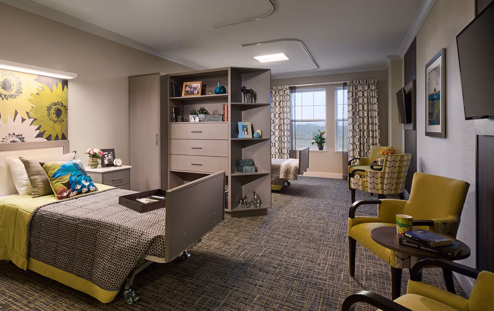 A spacious, well-lit bedroom in a senior living facility featuring two beds separated by a large shelving unit with drawers and decorative items. The room has patterned carpet, two yellow armchairs with a small round table between them, a wall-mounted TV, and large windows with patterned curtains allowing natural light to enter.