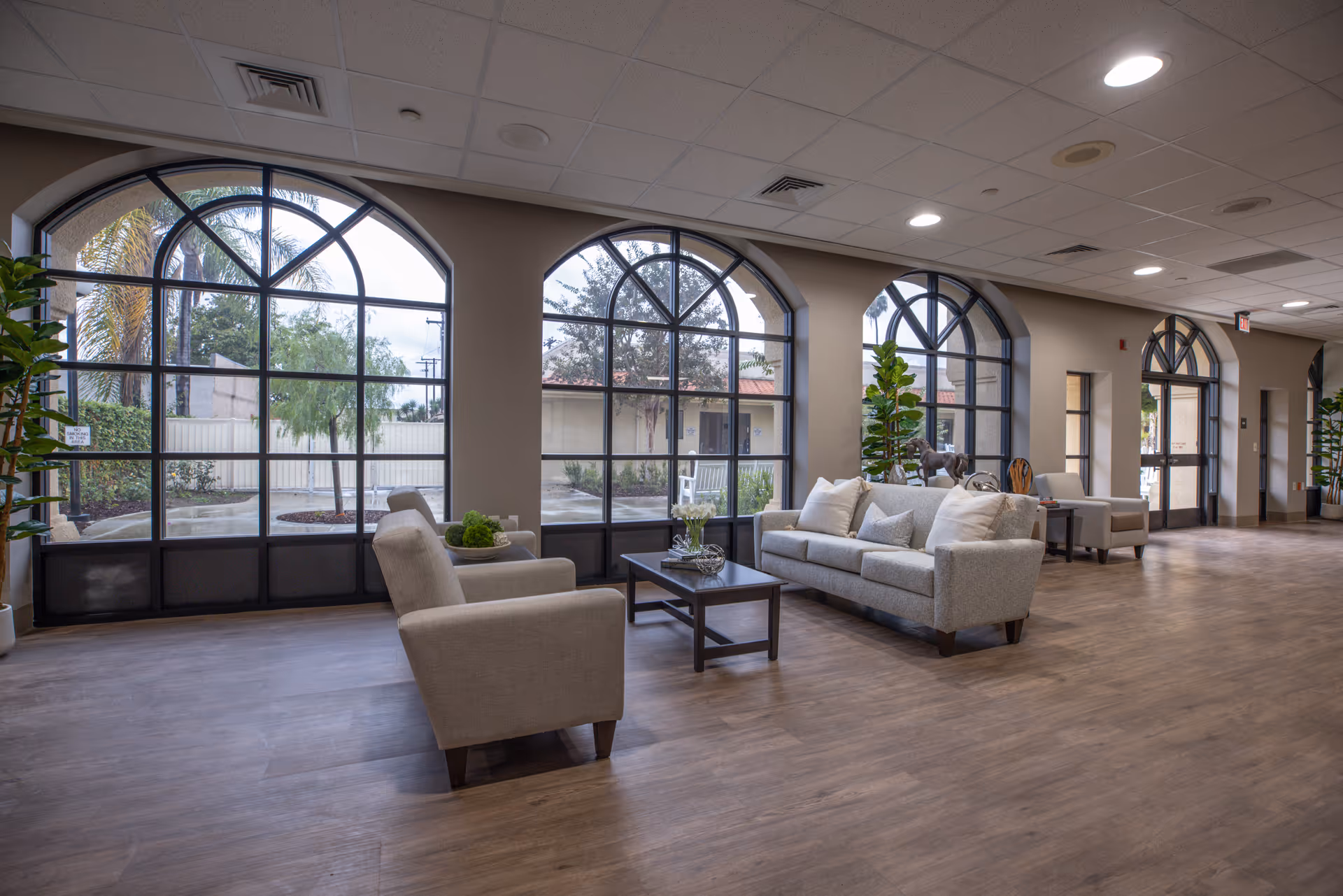 Bright senior living lobby with arched windows, beige seating, and potted plants.