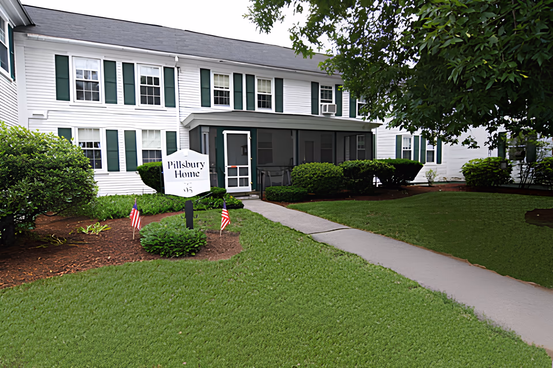 White two-story building with green shutters, a front lawn and a sign that reads 'Pillsbury Home' by the walkway.