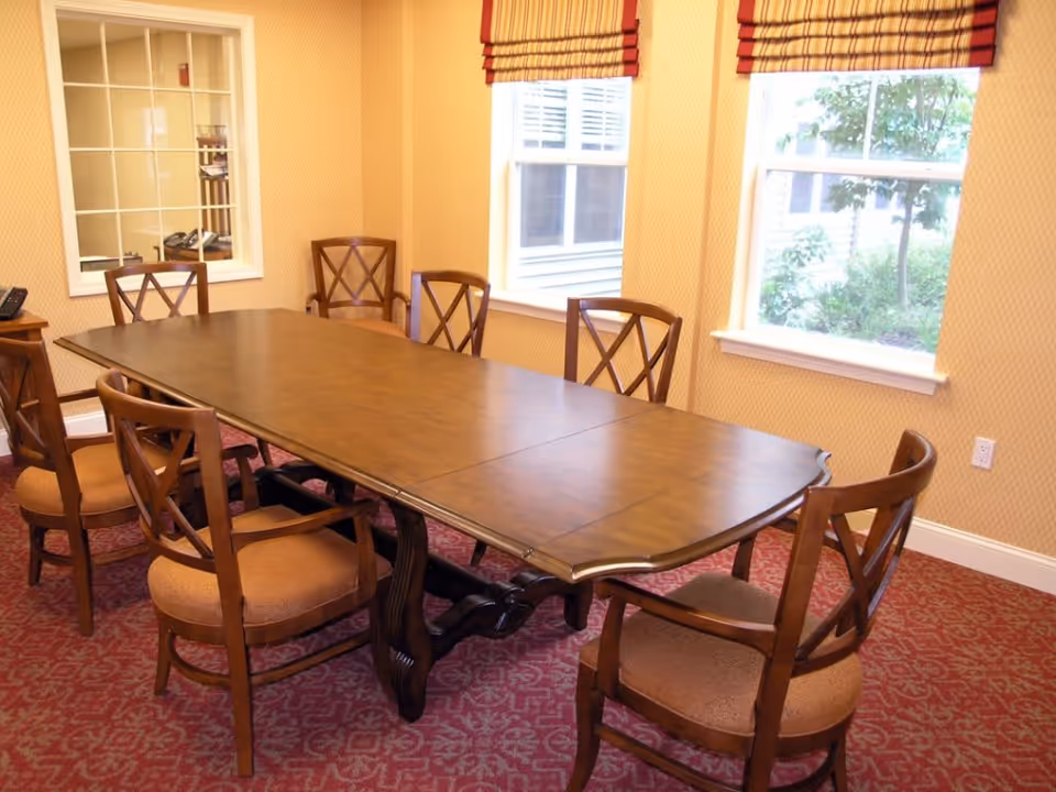 Wooden dining table surrounded by six chairs in a carpeted room with patterned wallpaper and two windows.