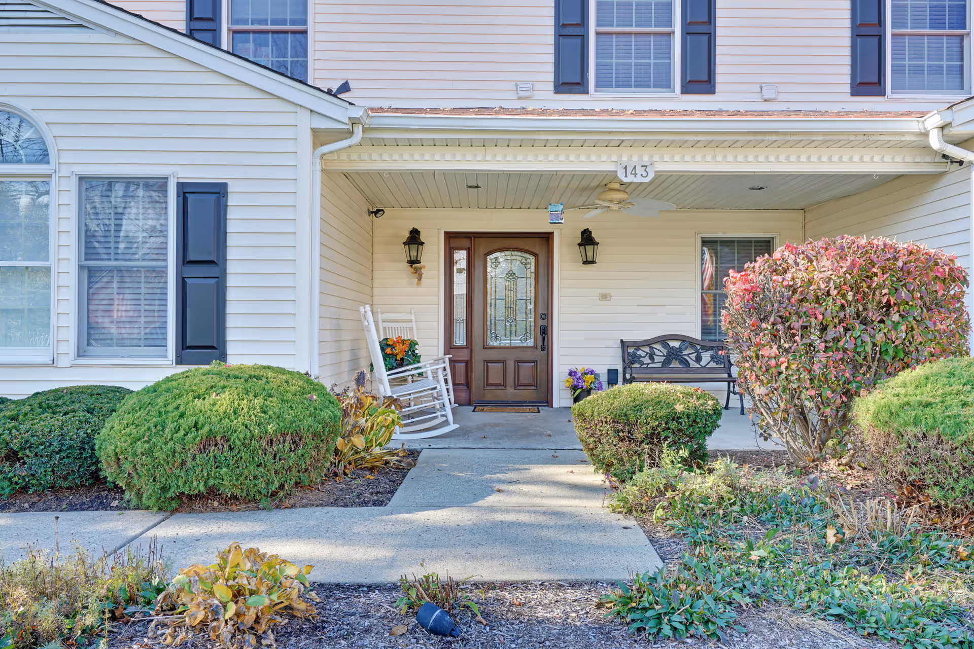 Front porch of a residential-style building with a wooden door featuring decorative glass panels. The porch has two black wall lanterns, a white rocking chair with a cushion and flowers, a black metal bench, and various bushes and plants in the garden beds surrounding the concrete walkway leading to the entrance.