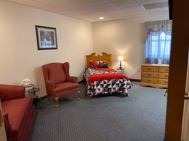 A bedroom in an assisted living facility featuring a single bed with a geometric patterned bedspread in red, black, and white. There is a wooden headboard, a bedside table with a lamp, a wooden dresser under a window with curtains, and two upholstered chairs in red. A framed picture hangs on the wall above one chair.