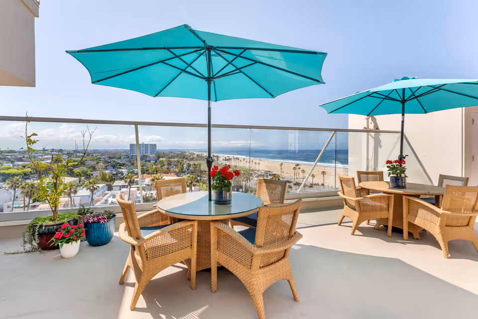 Outdoor balcony seating area with wicker tables and turquoise umbrellas overlooking the ocean.