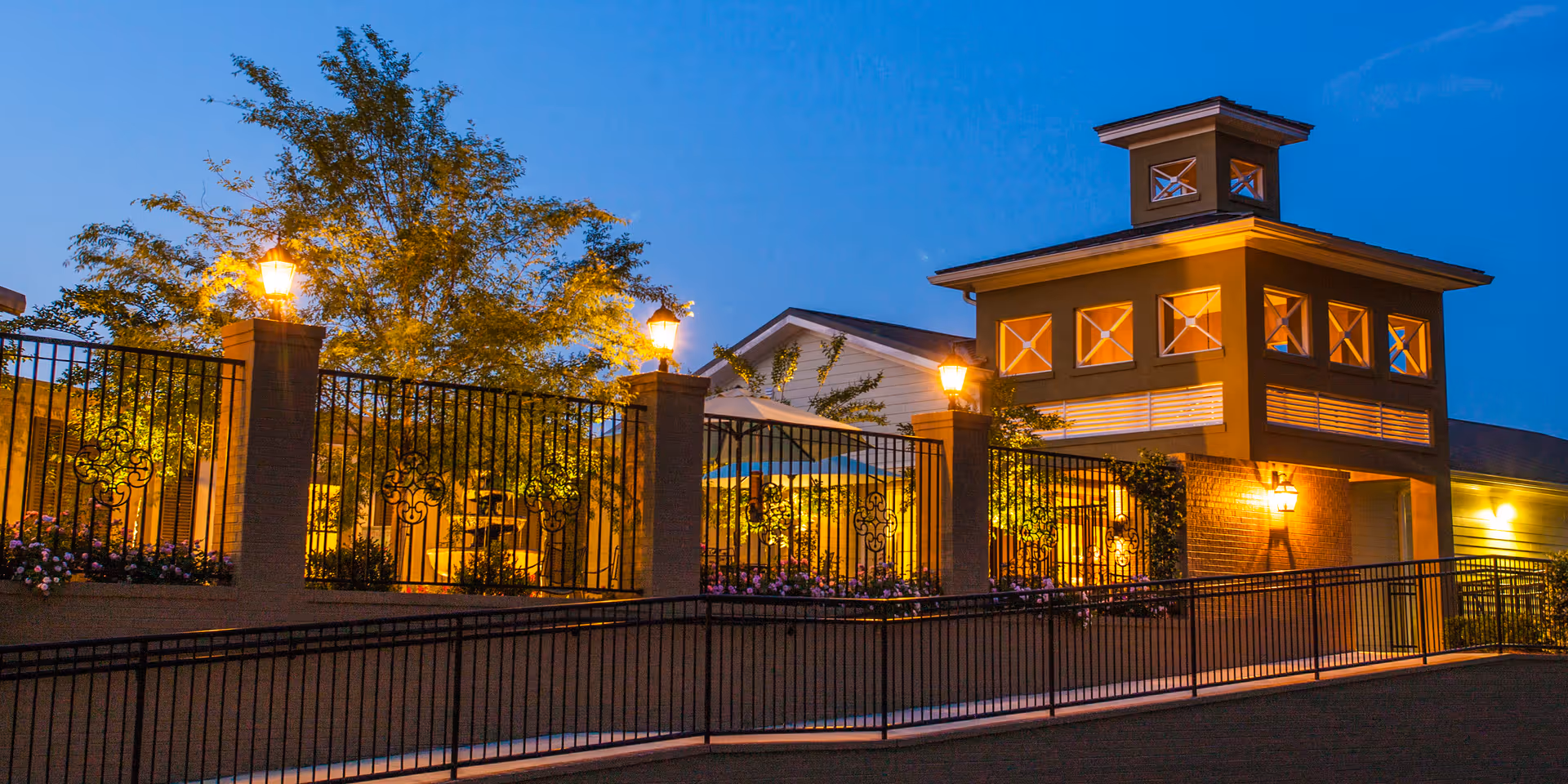 Evening view of an assisted living community exterior featuring a well-lit building with multiple windows and a decorative tower structure. The area is enclosed by a black wrought iron fence with brick pillars topped with lantern-style lights. Trees and plants are visible behind the fence, creating a warm and inviting atmosphere.