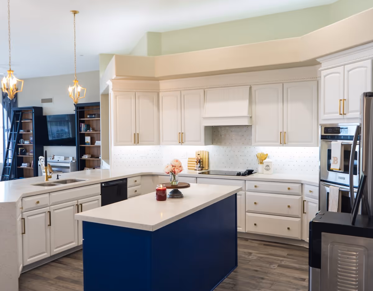 A modern kitchen with white cabinetry and gold handles, a blue island with a white countertop, a sink with a gold faucet, a built-in stovetop, and stainless steel double ovens. The kitchen opens into a living area with bookshelves, a TV, and pendant lights hanging from the ceiling.