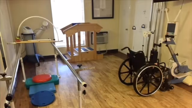 A rehabilitation room with parallel bars for walking exercises, a wheelchair, exercise equipment including a stationary bike, and a wooden step platform. The room has wood flooring and light-colored walls with a window and framed documents on the wall.