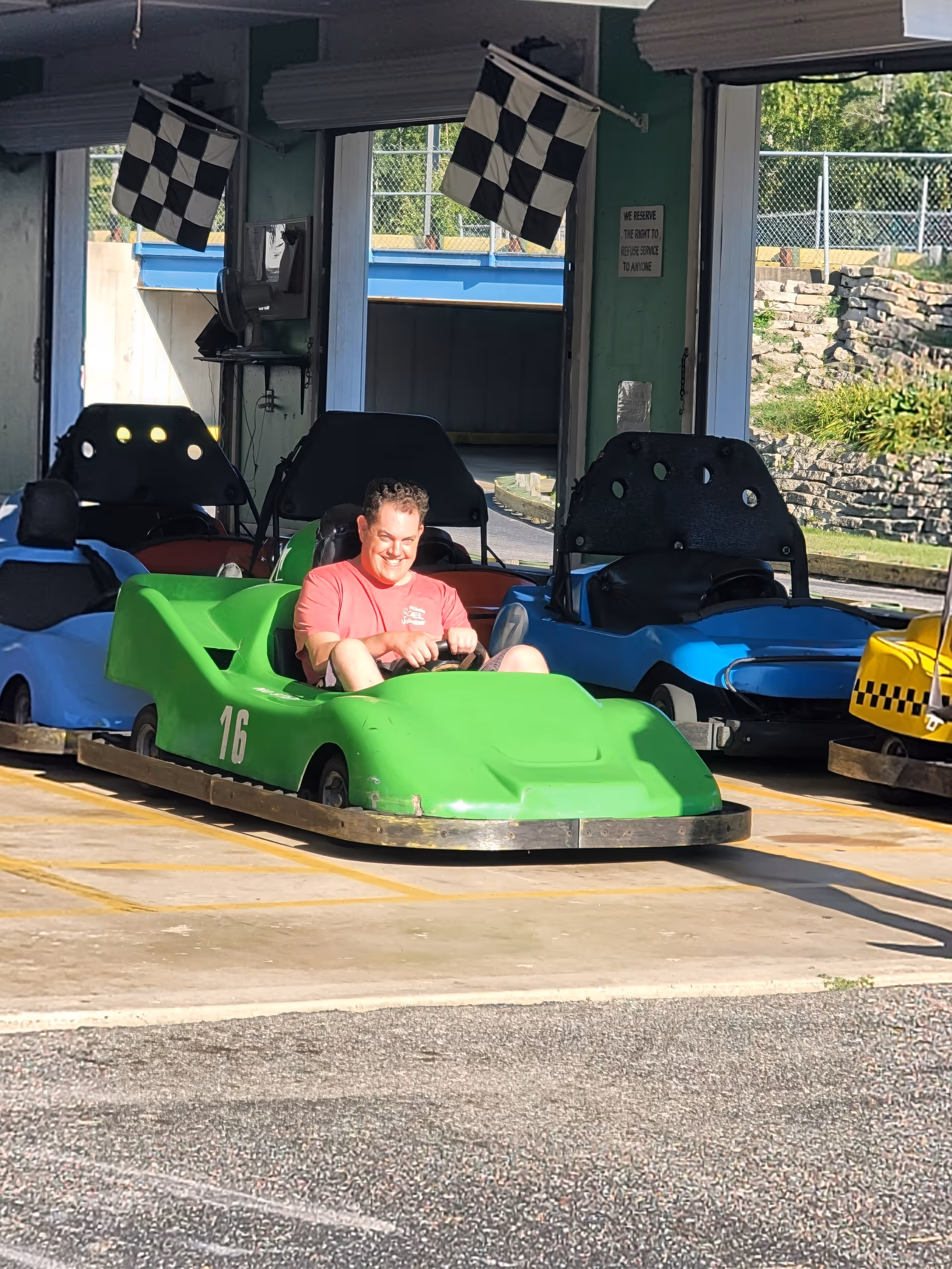 Man in pink shirt drives a bright green go-kart at outdoor track.