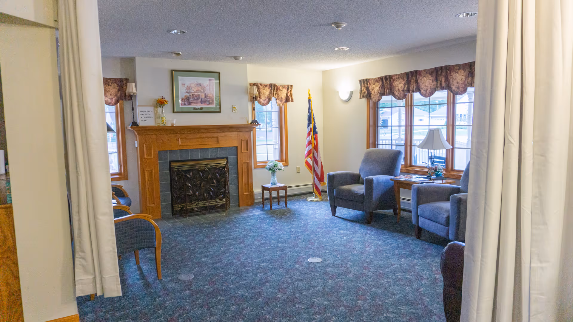 A cozy living room area with a wooden fireplace, two windows with floral valances, two gray armchairs, a small wooden table with a lamp, and an American flag in the corner. The room has carpeted flooring and light-colored walls.