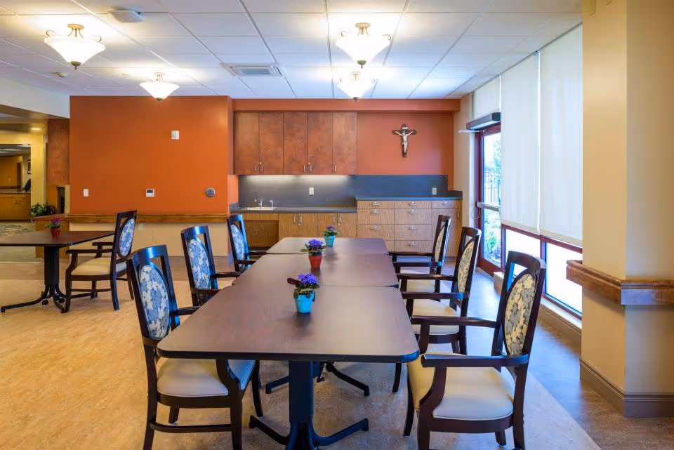 Spacious dining room with long tables, multiple chairs, small potted plants, and kitchenette.