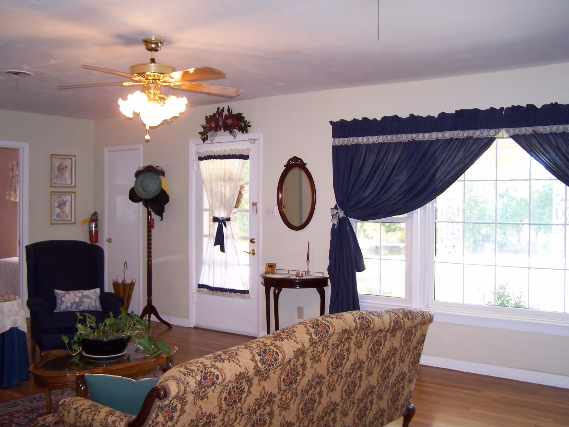 A cozy living room with a floral patterned sofa in the foreground, a wooden coffee table with a potted plant, a dark blue armchair with a decorative pillow, and a wooden hat stand with several hats. The room features a ceiling fan with lights, a small wooden table with a candle and a small picture frame, an oval mirror on the wall, and large windows with dark blue curtains letting in natural light. There is also a door with a sheer curtain tied with a dark blue ribbon.