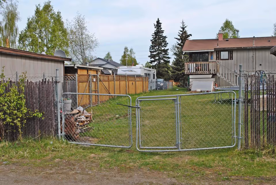 A fenced backyard area with a metal chain-link gate in the foreground. The yard has green grass and is bordered by wooden fences and a building with a balcony and stairs on the right side. Trees and a camper trailer are visible in the background under a cloudy sky.