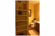 Narrow bathroom with left wooden shelves, vanity, and toilet at back under warm lighting.