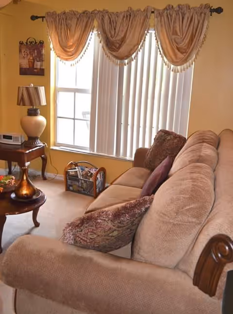 Cozy living room with a beige upholstered sofa and patterned pillows, coffee and side tables, a lamp, and a window with drapes and vertical blinds.