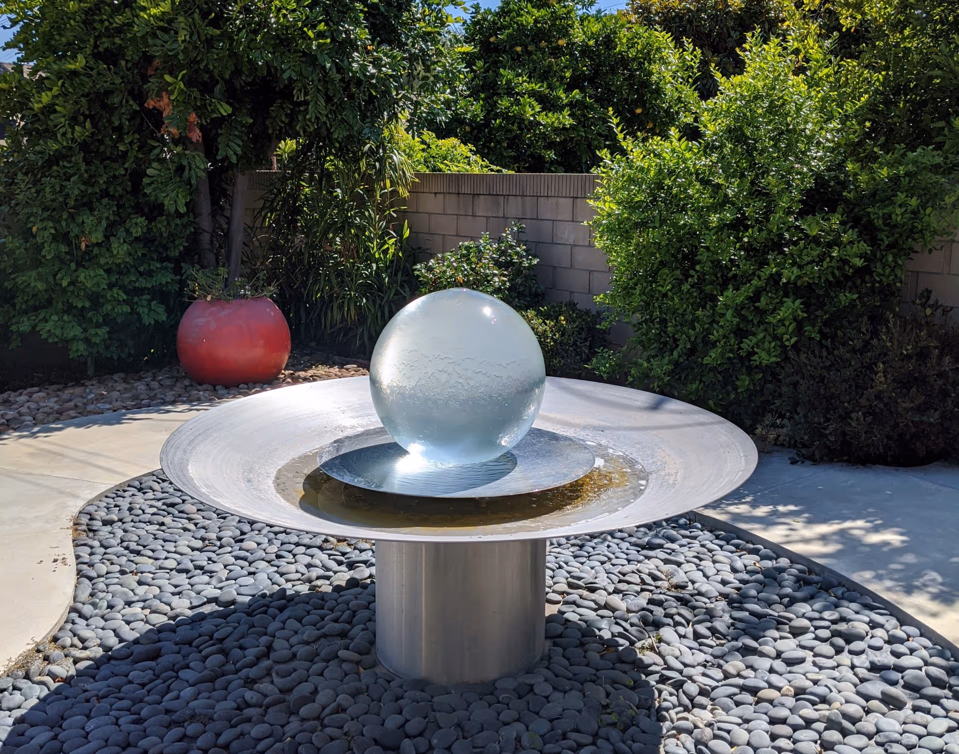 A modern outdoor water fountain featuring a large clear glass sphere resting on a metallic circular base, surrounded by smooth gray pebbles. The fountain is set in a garden area with lush green bushes and a red planter in the background, along with a concrete pathway.