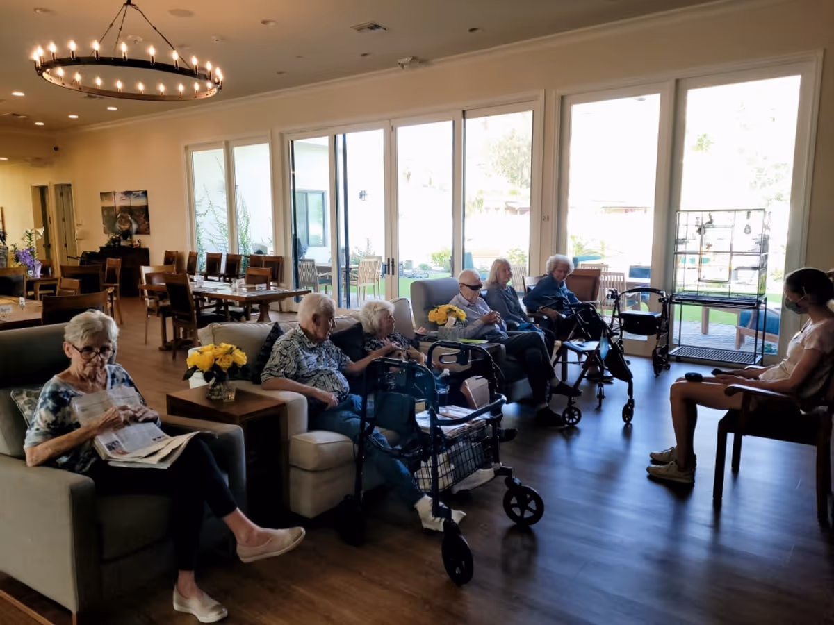 A group of elderly people sitting in a well-lit common area with large windows and sliding glass doors leading outside. Some are seated on sofas and chairs, with walkers nearby. A caregiver wearing a mask is seated on a chair facing them. The room has wooden floors, a chandelier, and tables with chairs in the background.