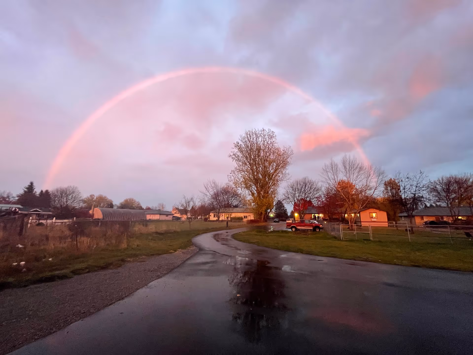 A rainbow arches over a wet residential street with trees and houses.
