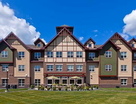 Three-story senior living building with peaked roofs, tan and green siding, and a well-maintained lawn.
