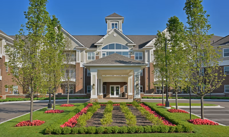 Front exterior view of Rose Senior Living Providence Park building with a covered entrance, neatly landscaped garden with rows of green shrubs and red flowers, and several young trees under a clear blue sky.