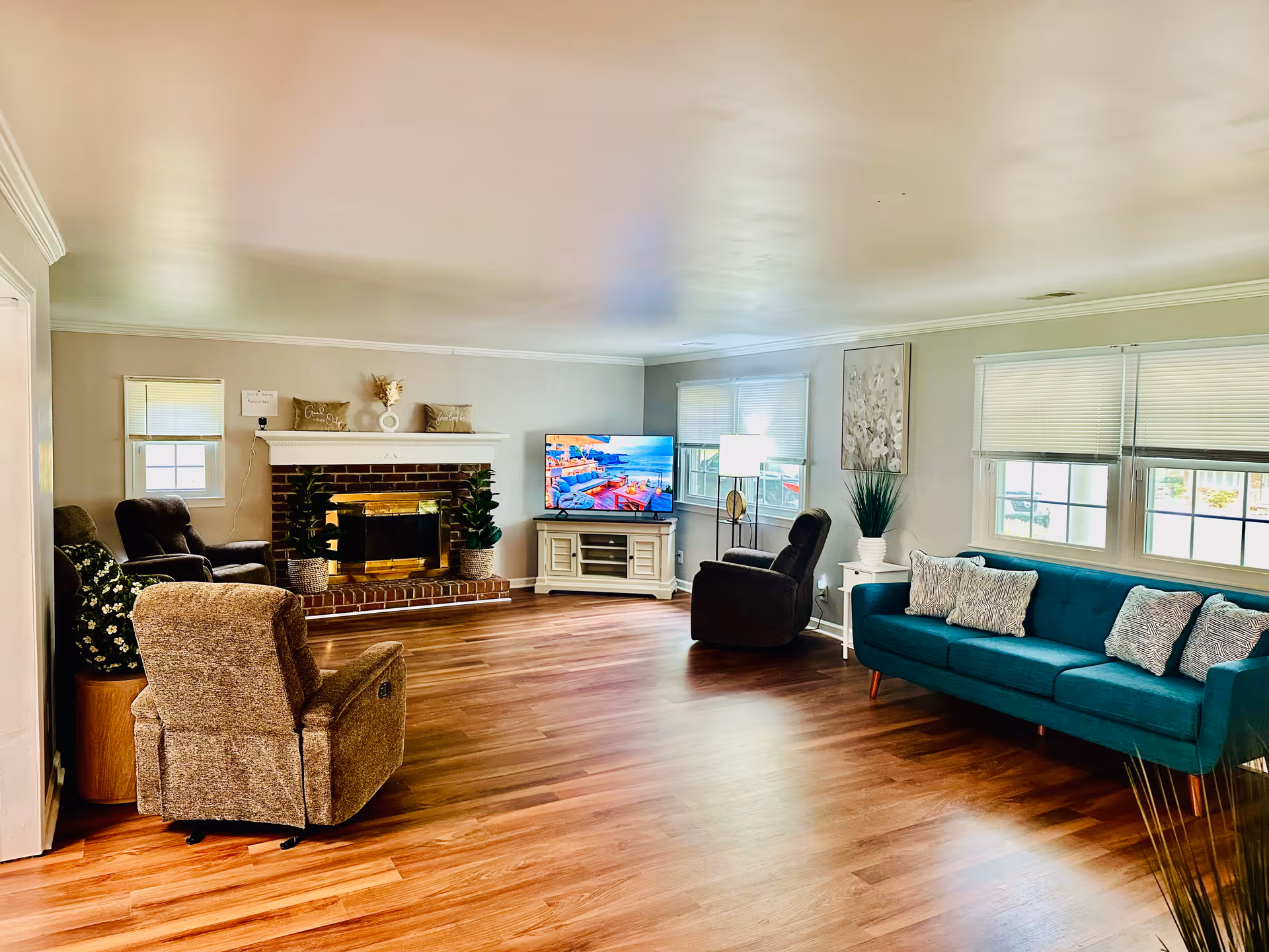 A spacious living room with wooden flooring, featuring a teal sofa with three cushions on the right, two brown recliner chairs on the left, and another recliner chair near a window. There is a brick fireplace with a white mantle decorated with plants and pillows. A flat-screen TV is placed on a white TV stand near the fireplace, and a floor lamp is positioned between the TV and the sofa. The room has multiple windows with blinds partially drawn, allowing natural light to fill the space.