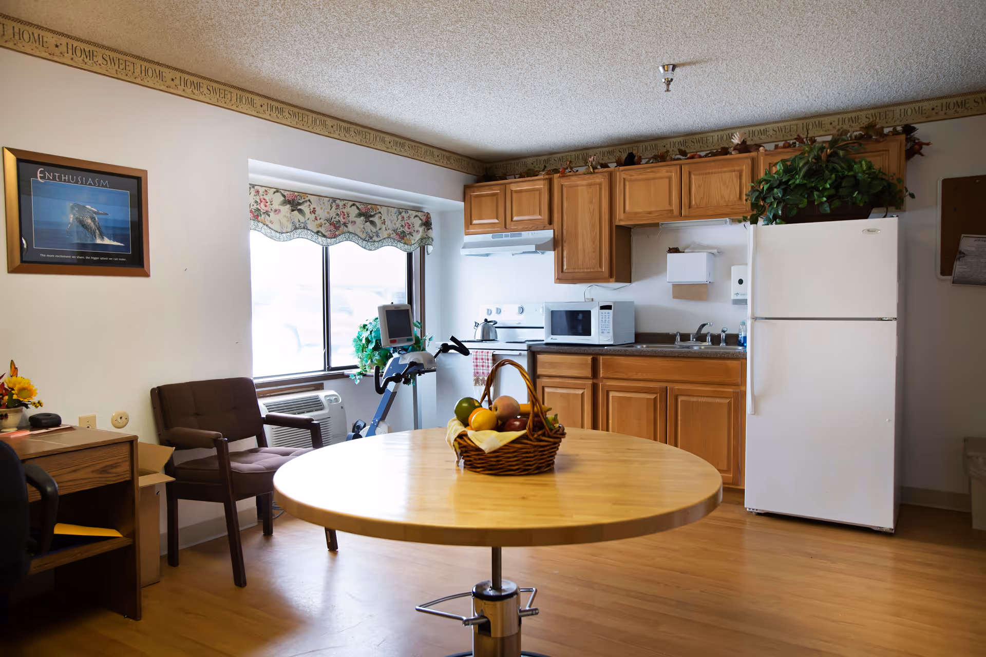 A cozy kitchen and dining area in a senior living facility with wooden cabinets, a white refrigerator, microwave, stove, and sink. A round wooden table with a basket of fruit is in the foreground. There is a window with floral valance letting in natural light, a brown cushioned chair, a small desk with a computer, and an exercise bike near the window. The walls have a decorative border with the phrase 'Home Sweet Home'.