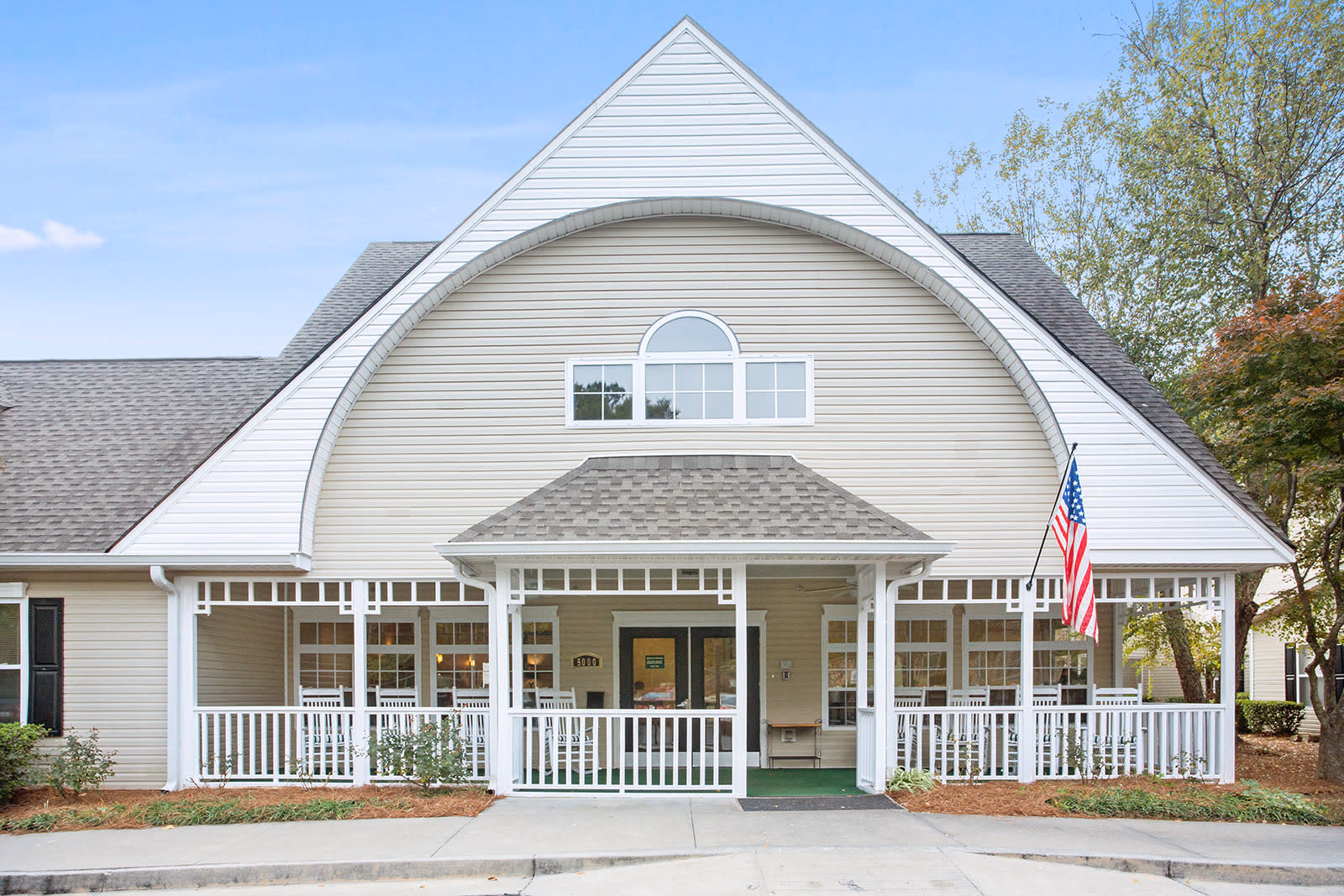Front exterior view of Governors Glen facility showing a beige building with white trim, a covered porch with white railings and rocking chairs, an American flag on the right side, and a clear blue sky in the background.