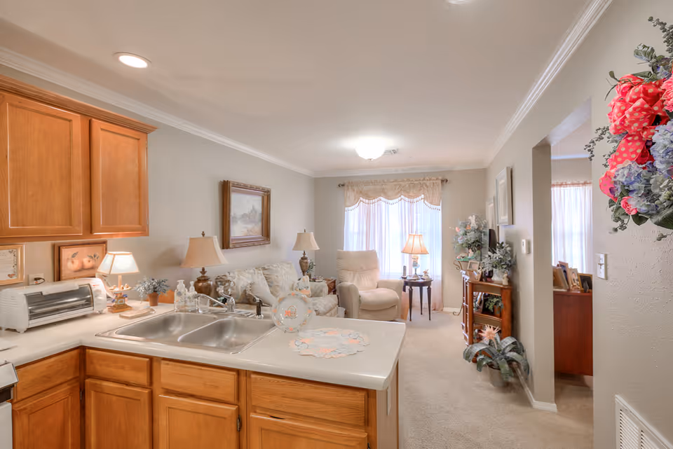 View of a cozy living area and kitchen in a senior living facility. The kitchen features wooden cabinets, a double sink, and a countertop with decorative items. The living room has a beige sofa, an armchair, side tables with lamps, a TV on a wooden stand, and large windows with sheer curtains allowing natural light to fill the space. Floral decorations are visible on the wall and furniture.