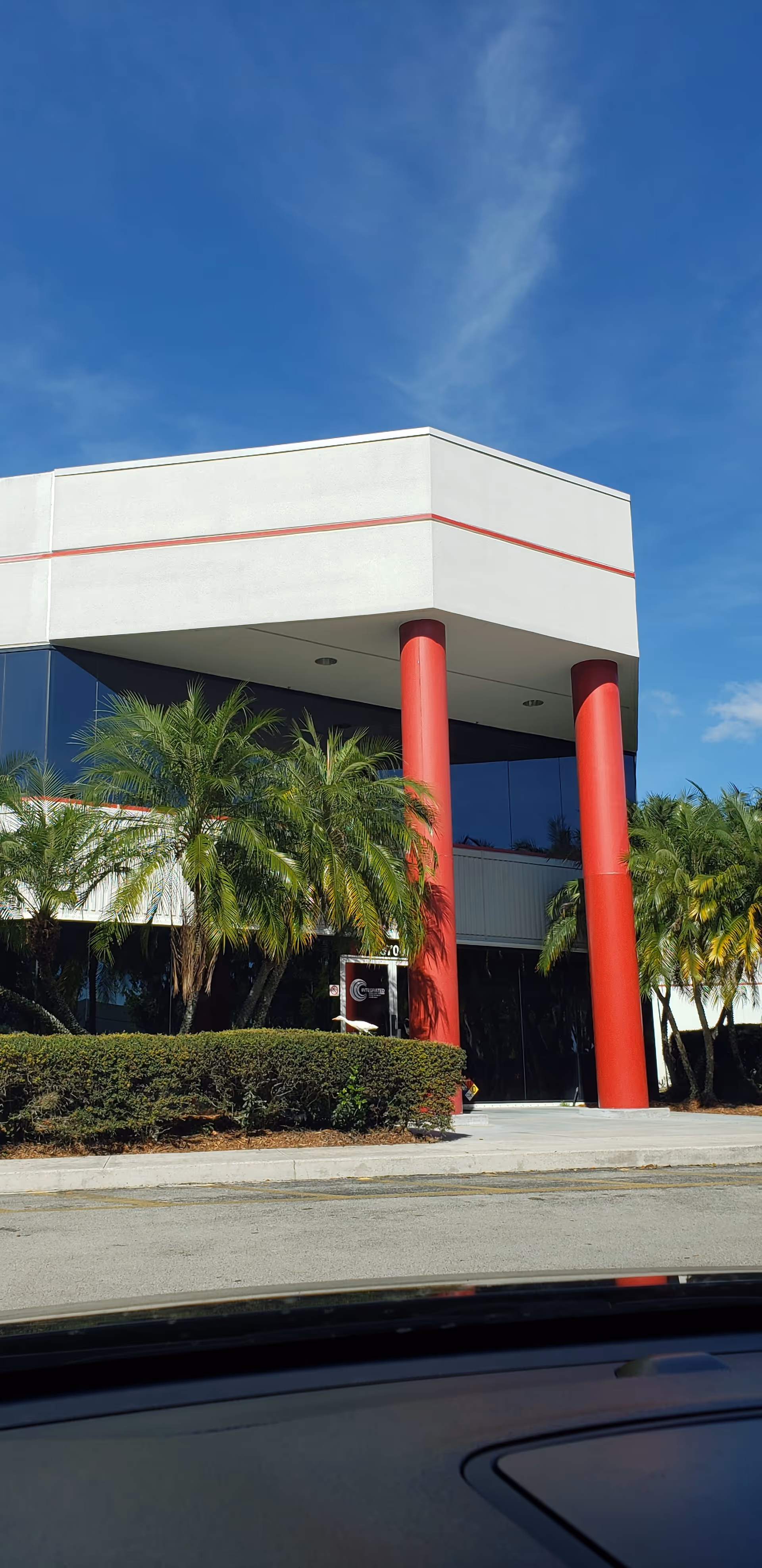 Modern building exterior with red columns, palm trees, and a clear blue sky.