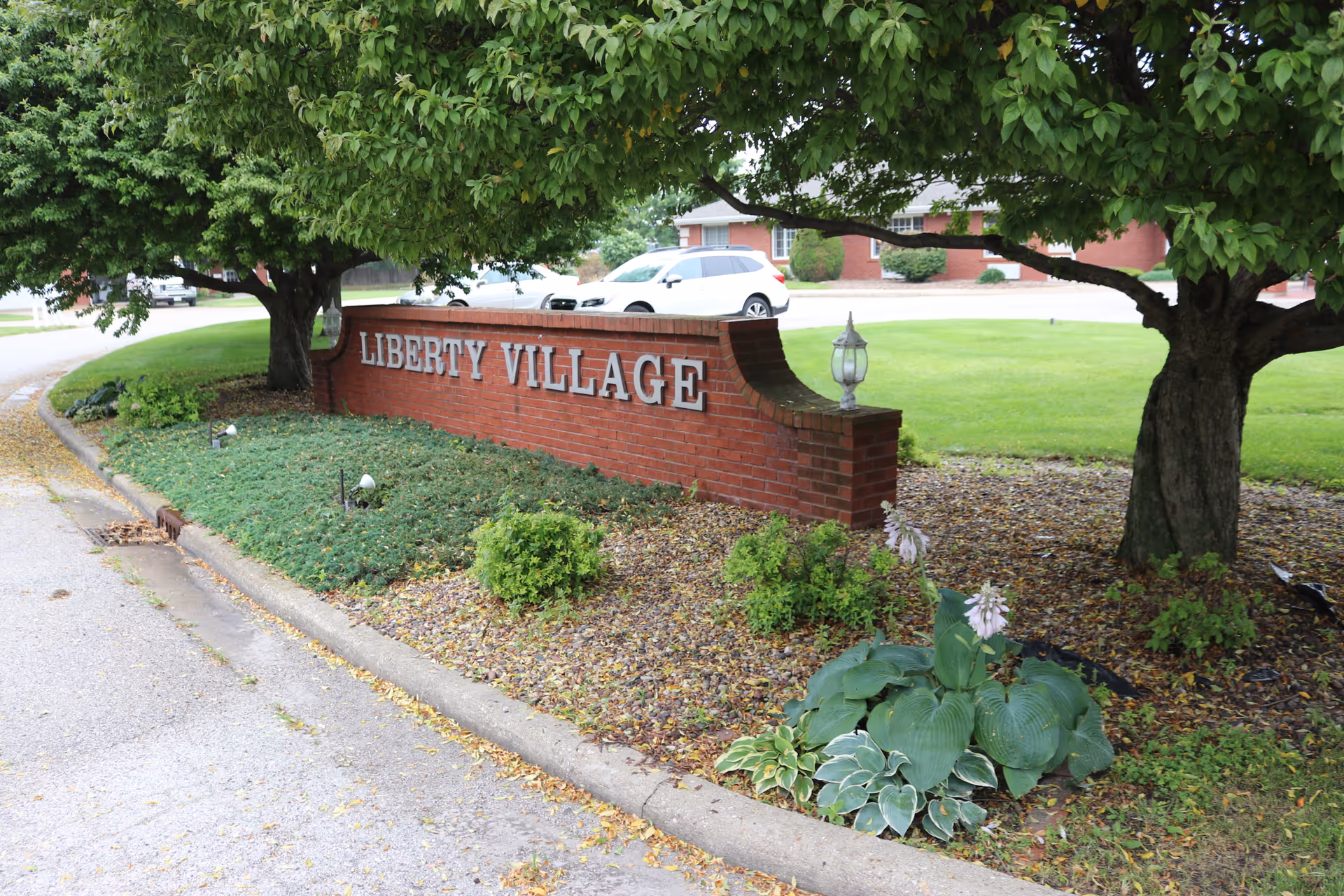 A landscaped entrance sign for Liberty Village made of red brick with silver letters. The sign is surrounded by green shrubs, plants, and trees, with a grassy lawn and a white car in the background.