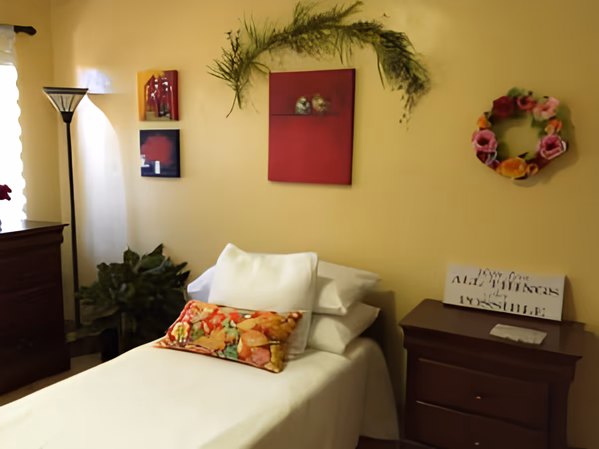 A cozy bedroom with a single bed covered in white sheets and pillows, including a colorful decorative pillow. The walls are painted yellow and decorated with a red painting, two smaller artworks, a green leafy garland, and a floral wreath. There is a wooden dresser on the left and a wooden nightstand on the right with a small sign and a piece of paper on it. A tall floor lamp stands near the window with sheer white curtains, and a green plant is placed in the corner.