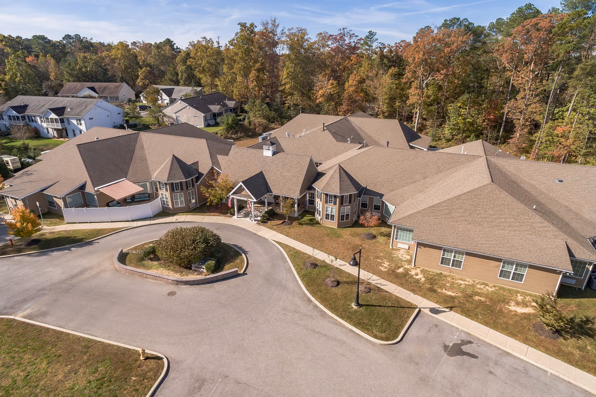 Aerial view of a single-story senior living facility with multiple connected buildings featuring beige siding and brown roofs, surrounded by trees with autumn foliage. There is a circular driveway with a landscaped island in front of the main entrance.