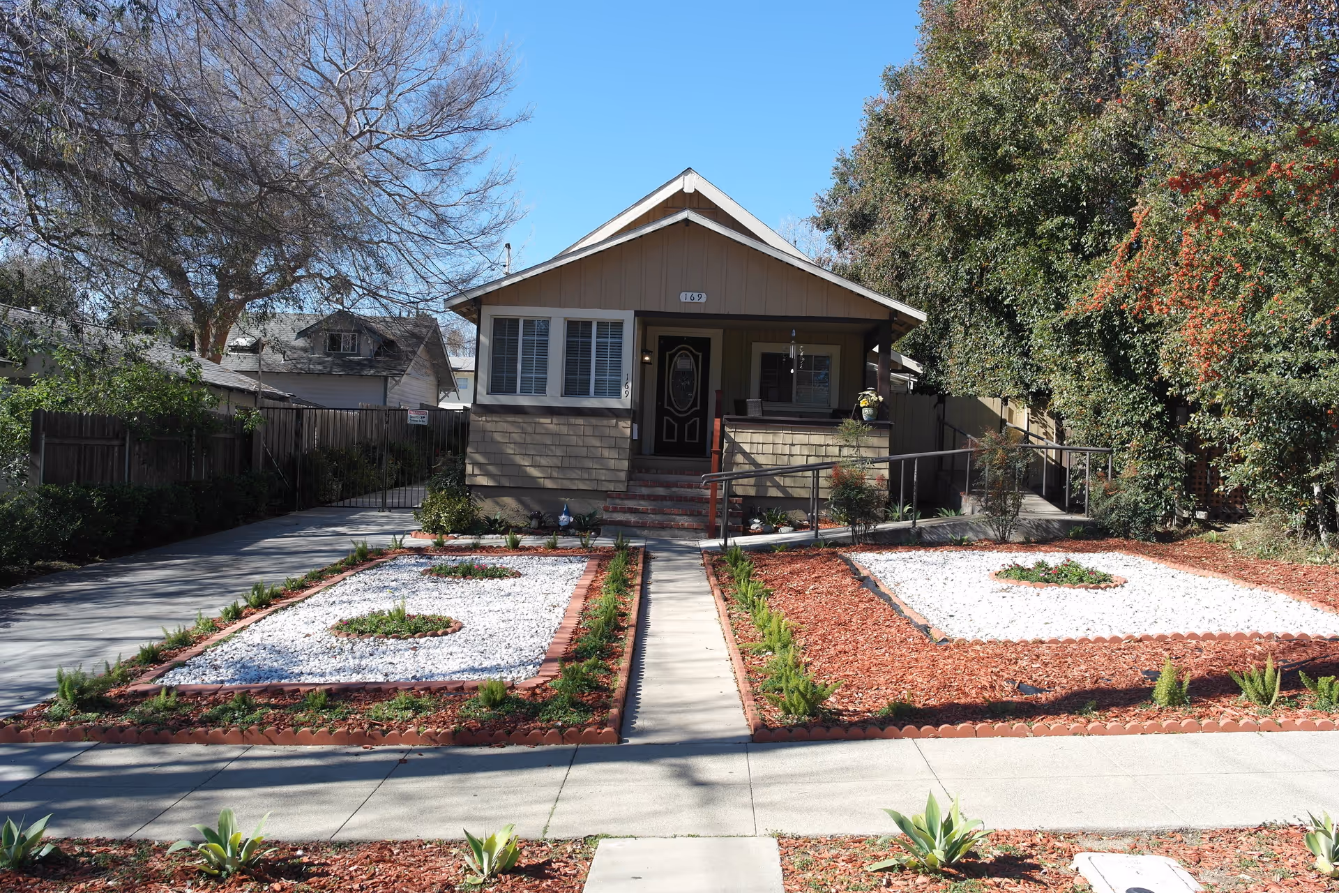 Front exterior view of a single-story house with a small porch, a black front door with decorative glass, and two large windows. The house number 169 is displayed above the door. The front yard is landscaped with two rectangular garden beds bordered with red bricks, filled with white rocks and mulch, and small plants. A concrete walkway leads from the sidewalk to the porch steps, and there is a wheelchair ramp on the right side. Trees and bushes surround the property under a clear blue sky.